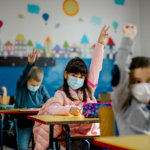 kids wearing mask in classroom raising hand