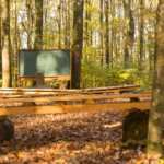 Empty outdoor class room in forest with blank classic green chalk board, school desks and benches for students with trees as backdrop and brown autumn colour leaves on ground. Picture meant to illustrate outdoor education and how it helps building positive youth-adult relationships.